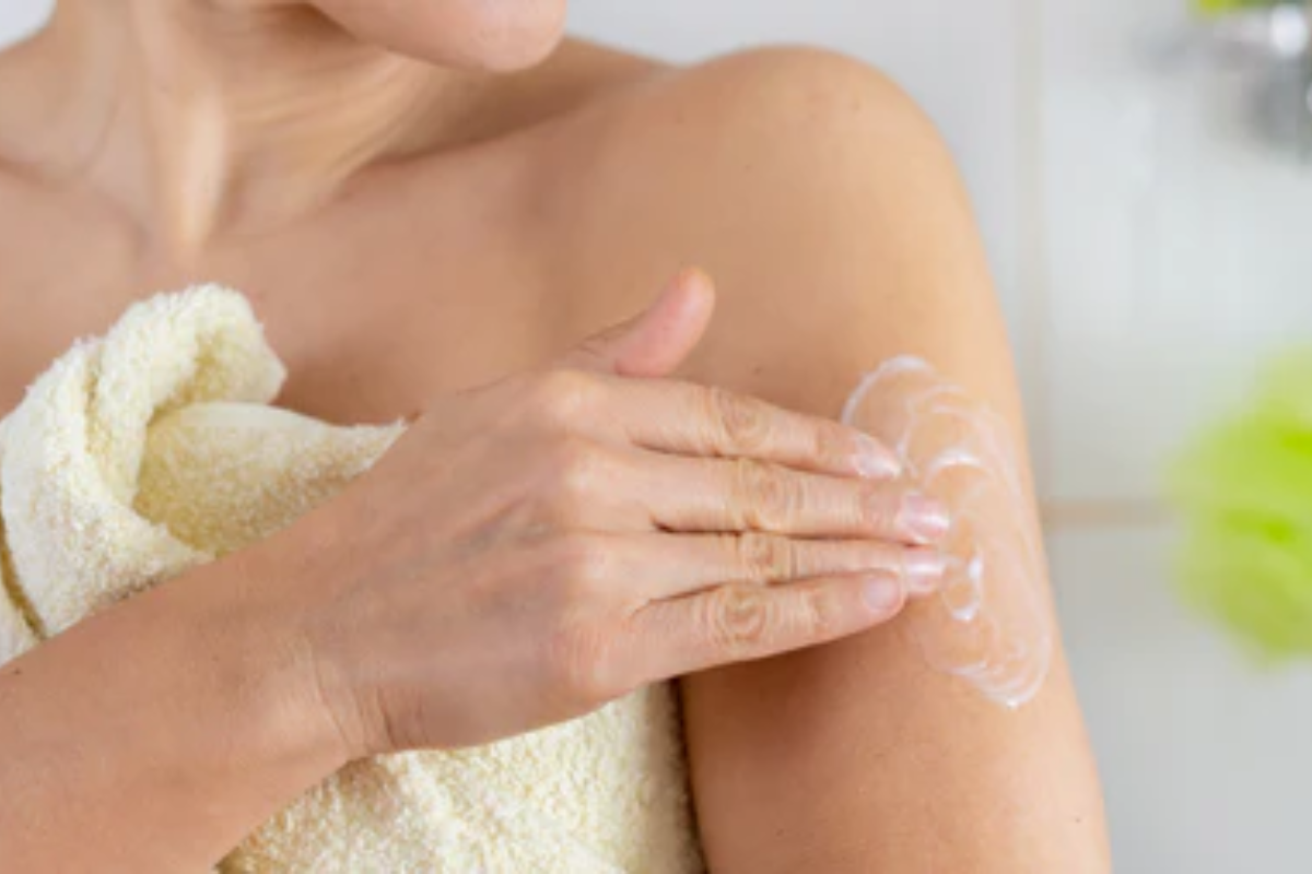 Midlife woman wrapped in a towel applying HRT cream to her upper arm after a shower.