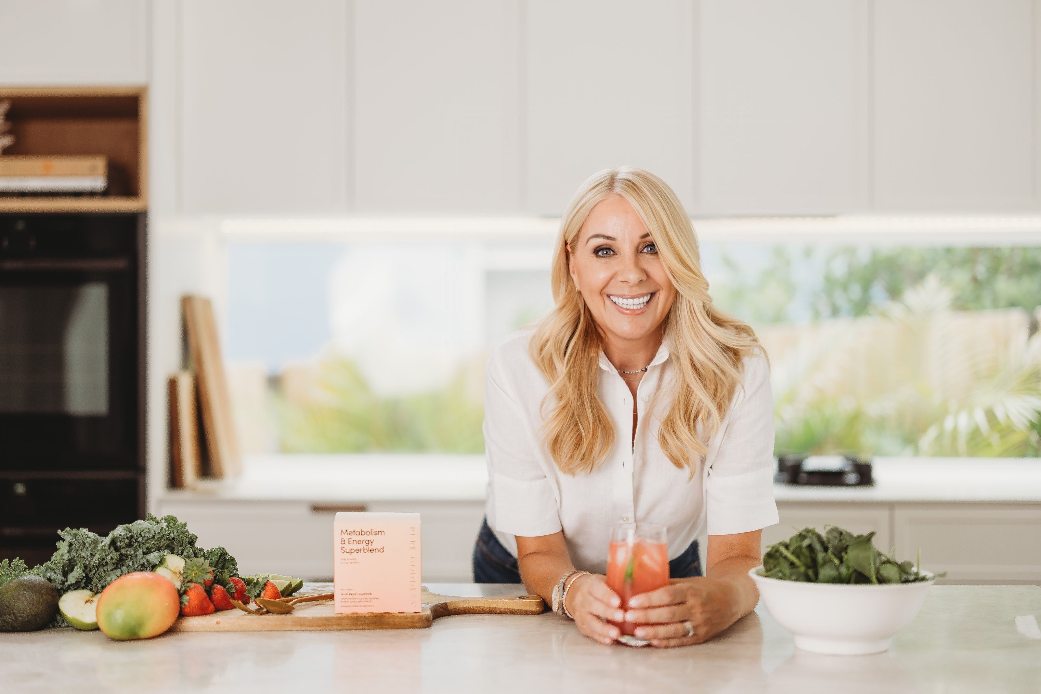 Smiling midlife woman in a bright kitchen holding a wellness drink, surrounded by leafy greens, fruit, and Pure Goddess Metabolism & Energy Superblend. promoting natural hormone support and healthy weight loss