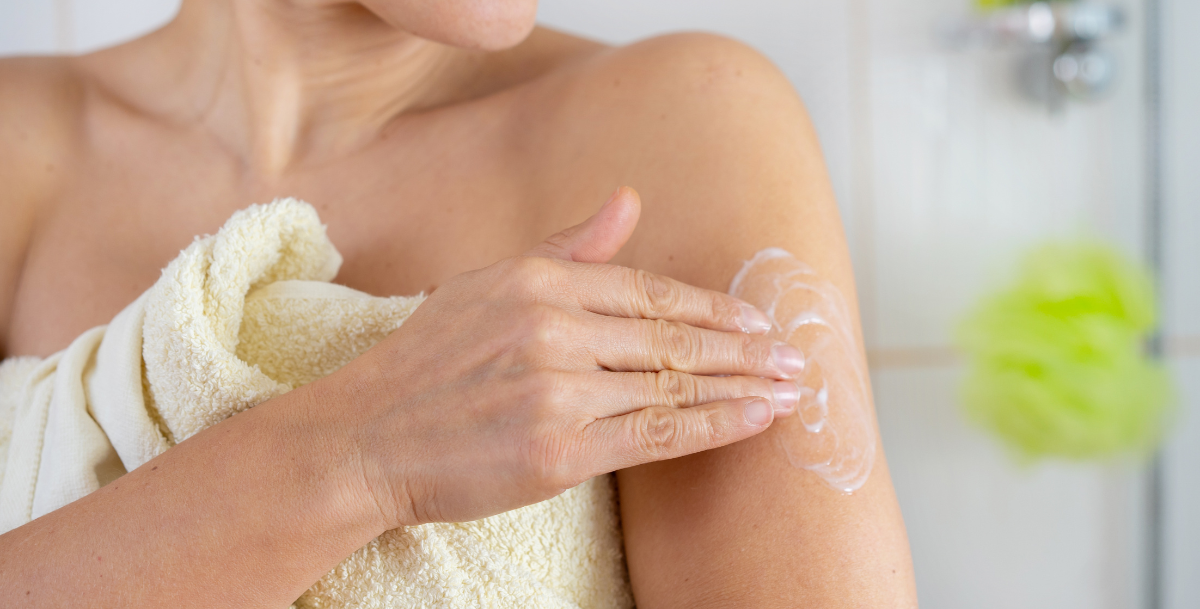 Midlife woman wrapped in a towel applying HRT cream to her upper arm after a shower.